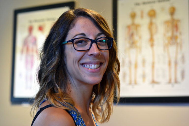 A woman in glasses is smiling in front of a wall of anatomy pictures.