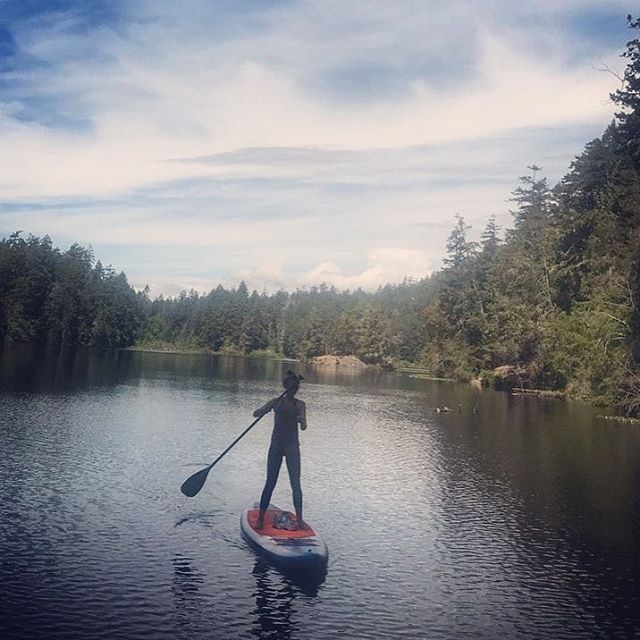 A woman is at the centre of the photo facing us on her paddle board. She is on a lake with large evergreen trees all along the shore.