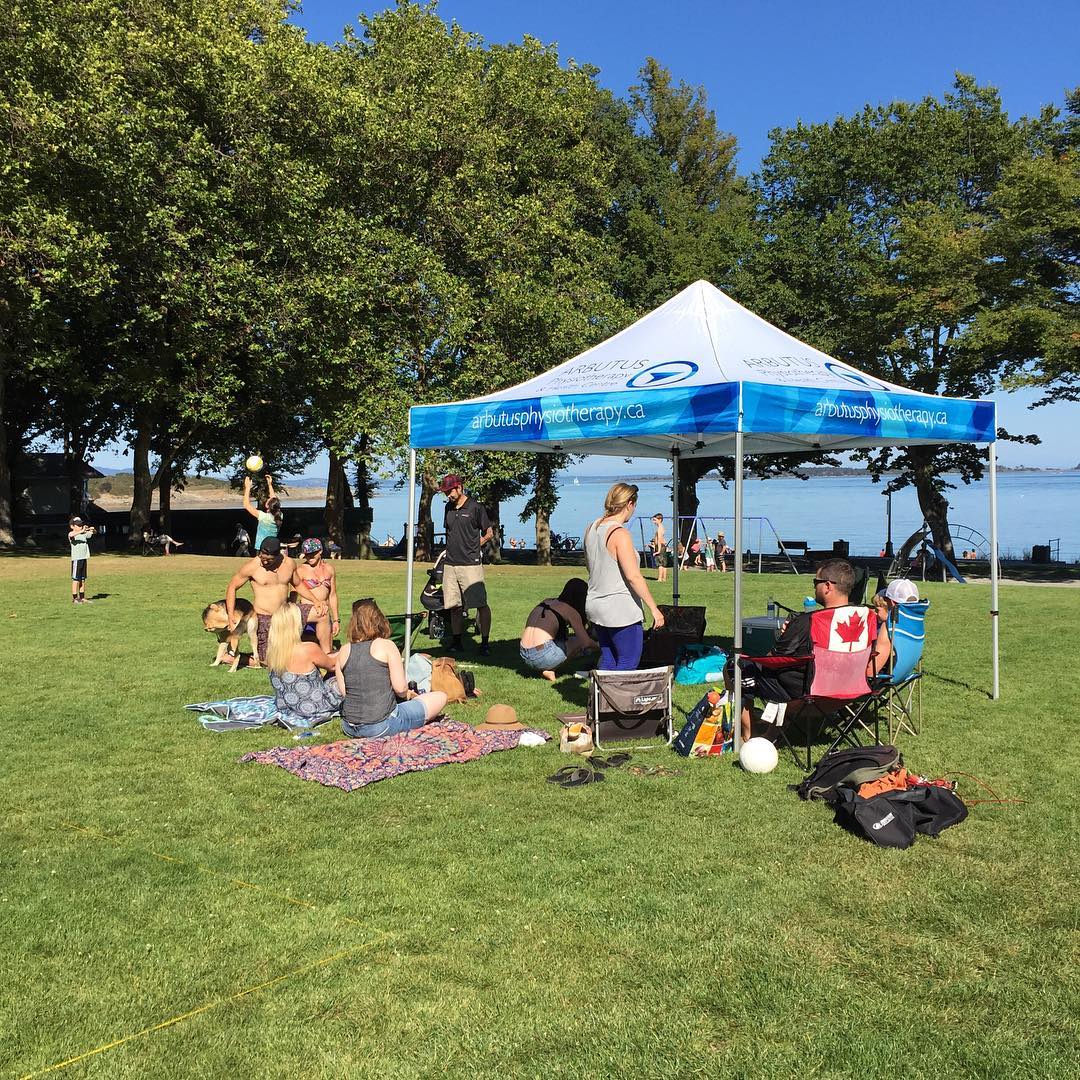 This is a photo of the arbutus team outside on the grass in front of Willows Beach. It is a sunny day, and they are under a white and light blue arbutus physio tent.