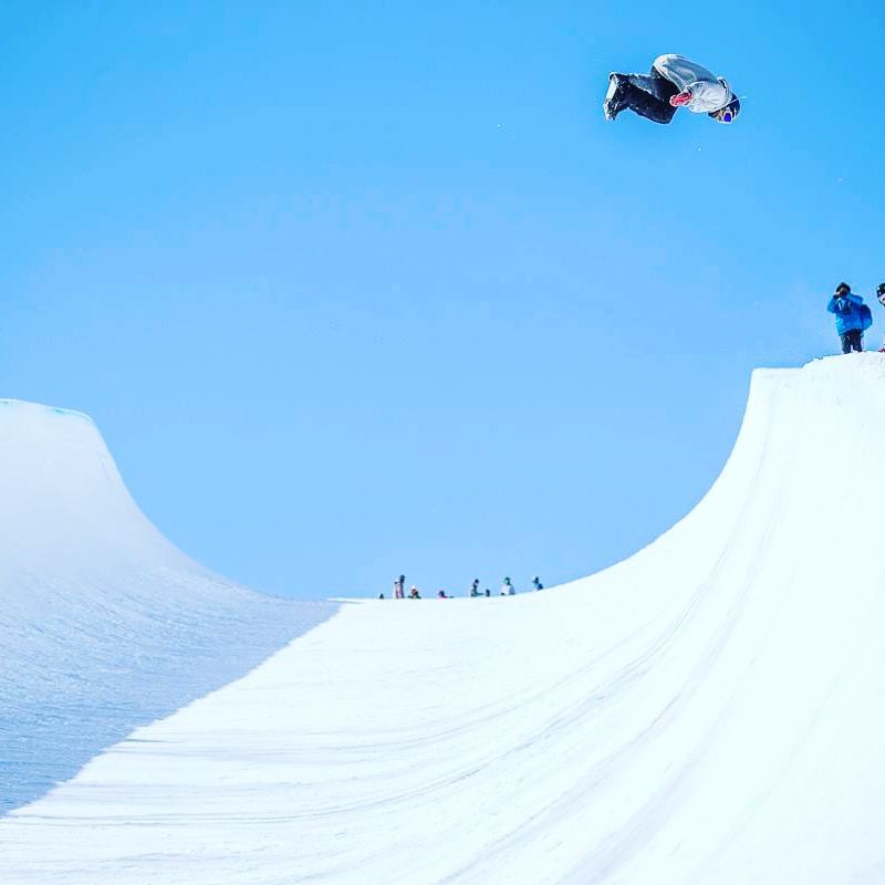 This is an image of Mike doing a jump on a snowboarding course. He is in the air on the right of the image and mid jump.