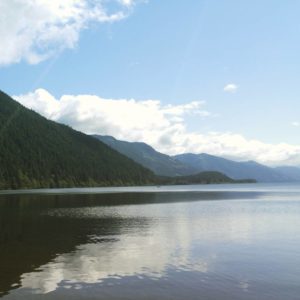 This is an image of Cowichan lake with clear, still water. The sky is blue with white fluffy clouds and been hills to the left.