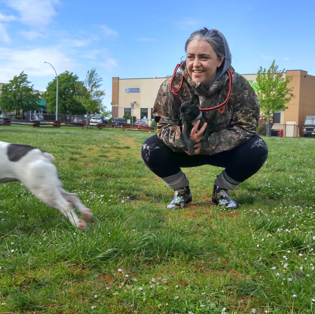 Smiling woman crouching on a field, holding a puppy and watching the dog in the foreground running off: by Save-on Foods and Starbucks.