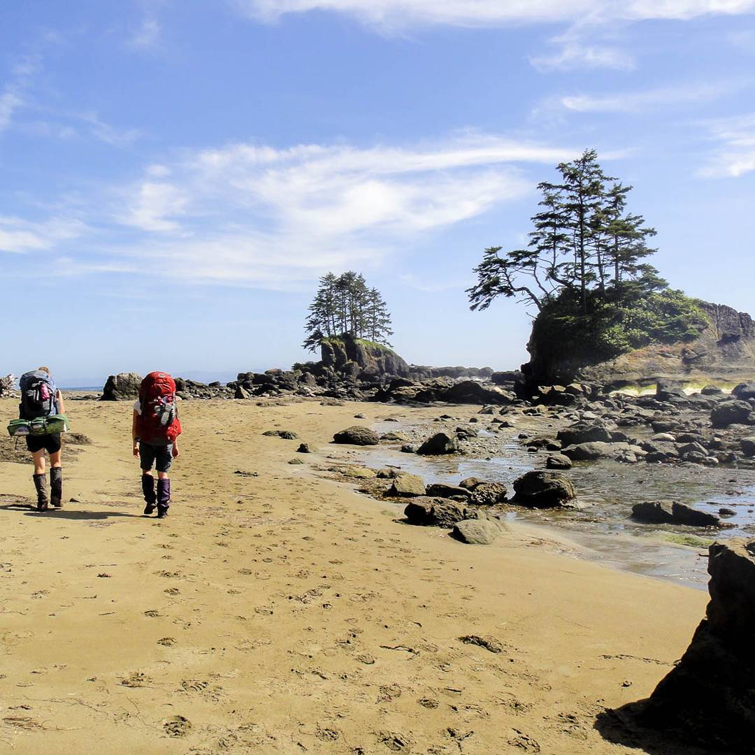 Two women hike along a sandy beach on the west coast trail, with stunning rock and tree formations along the shore.