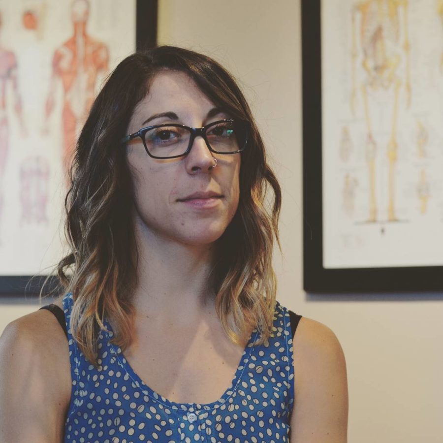 Ashley in the clinic with a neutral expression and anatomy drawing in the background.