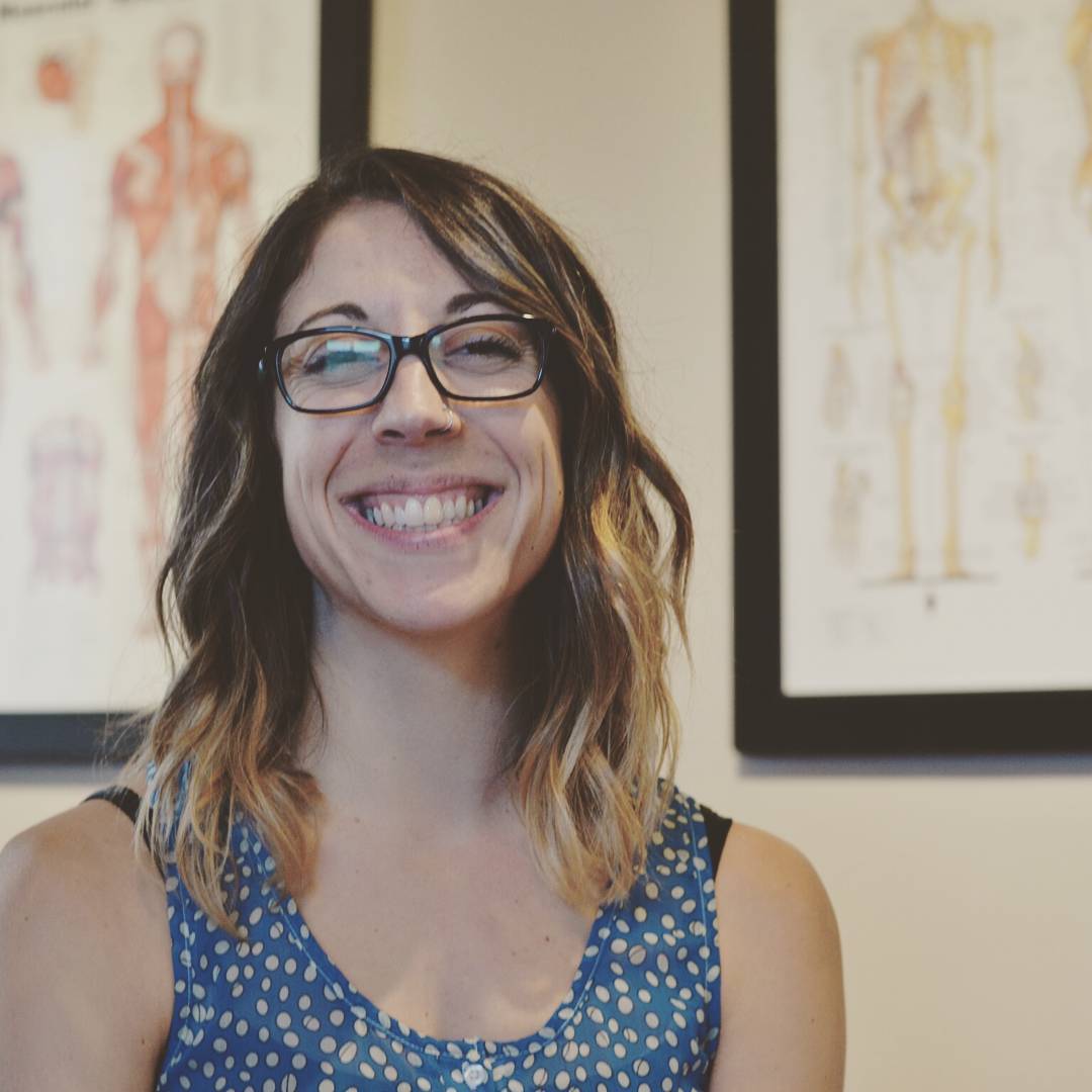 Ashley smiling brightly in the clinic with anatomy drawing in the background.