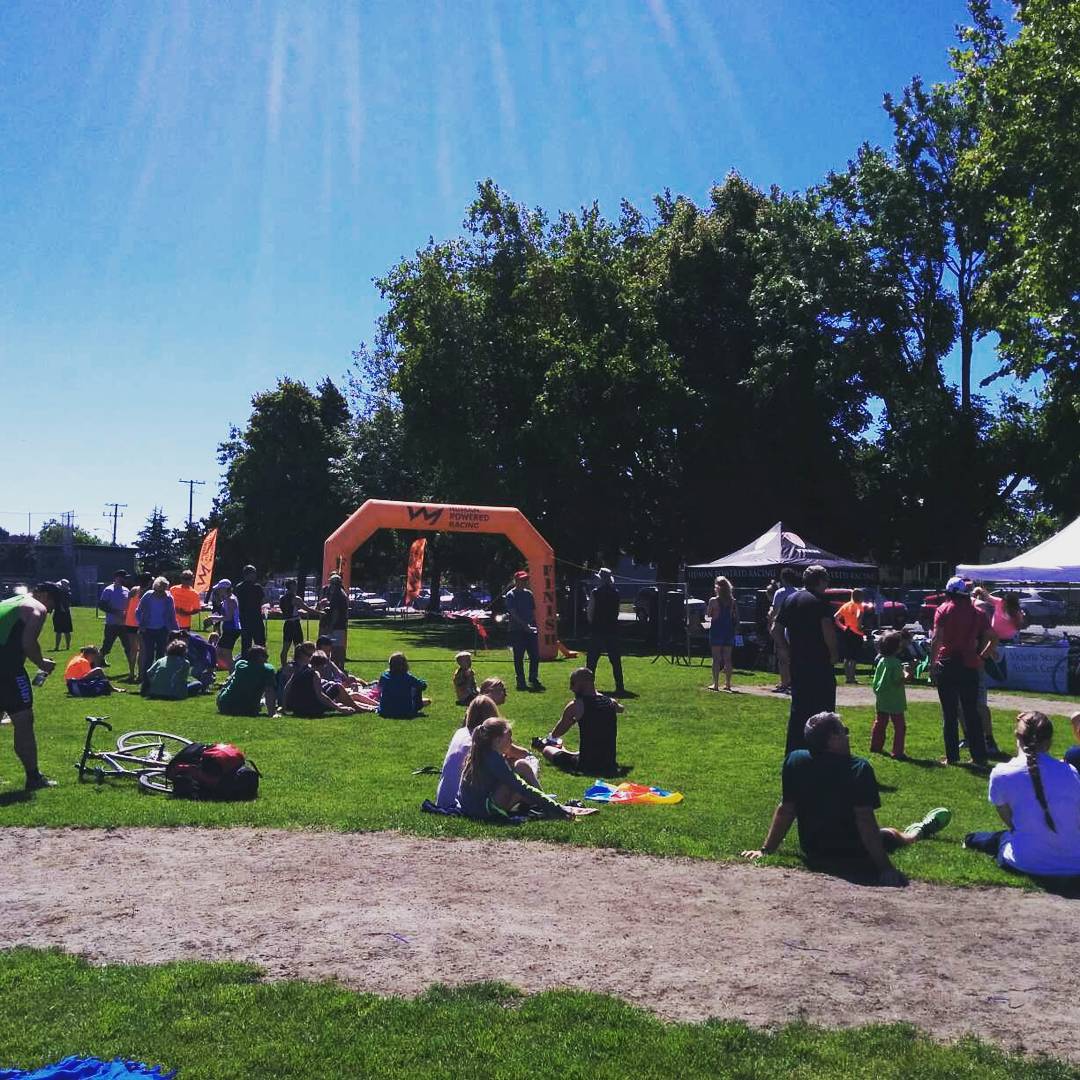 A sunny, grassy field with people sitting in many small groups, with a blow-up archway and branded tents in the background.