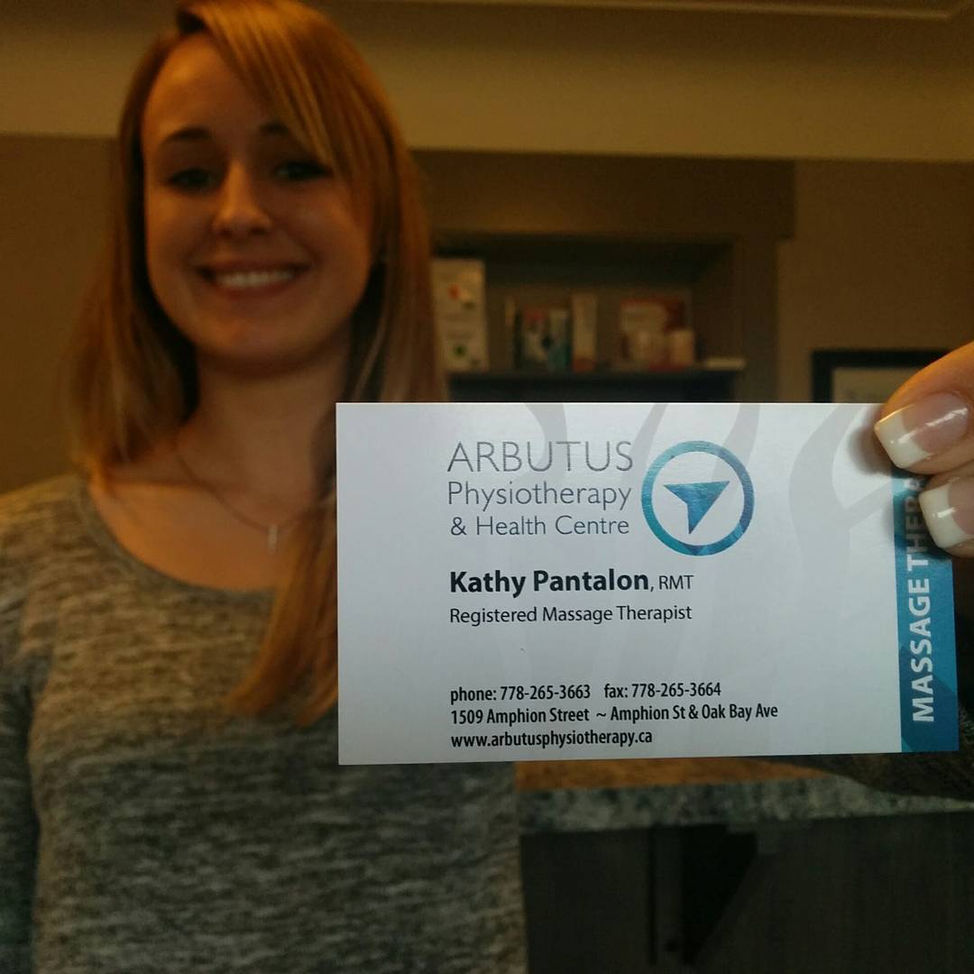 A blonde woman to the left it standing in a light brown office and smiling. She is out of focus. She is holding a card out in front of her to the right, which is in focus. It has the Arbutus logo of a blue arrow in a blue circle at the top and says "Kathy Pantalon, Recistered Massage Therapist, phone 778-265-3663, fax 778-265-3664. 1509 Amphion Street -- Amphion St. and Oak Bay Ave, www. arbutusphysiotherapy.ca."