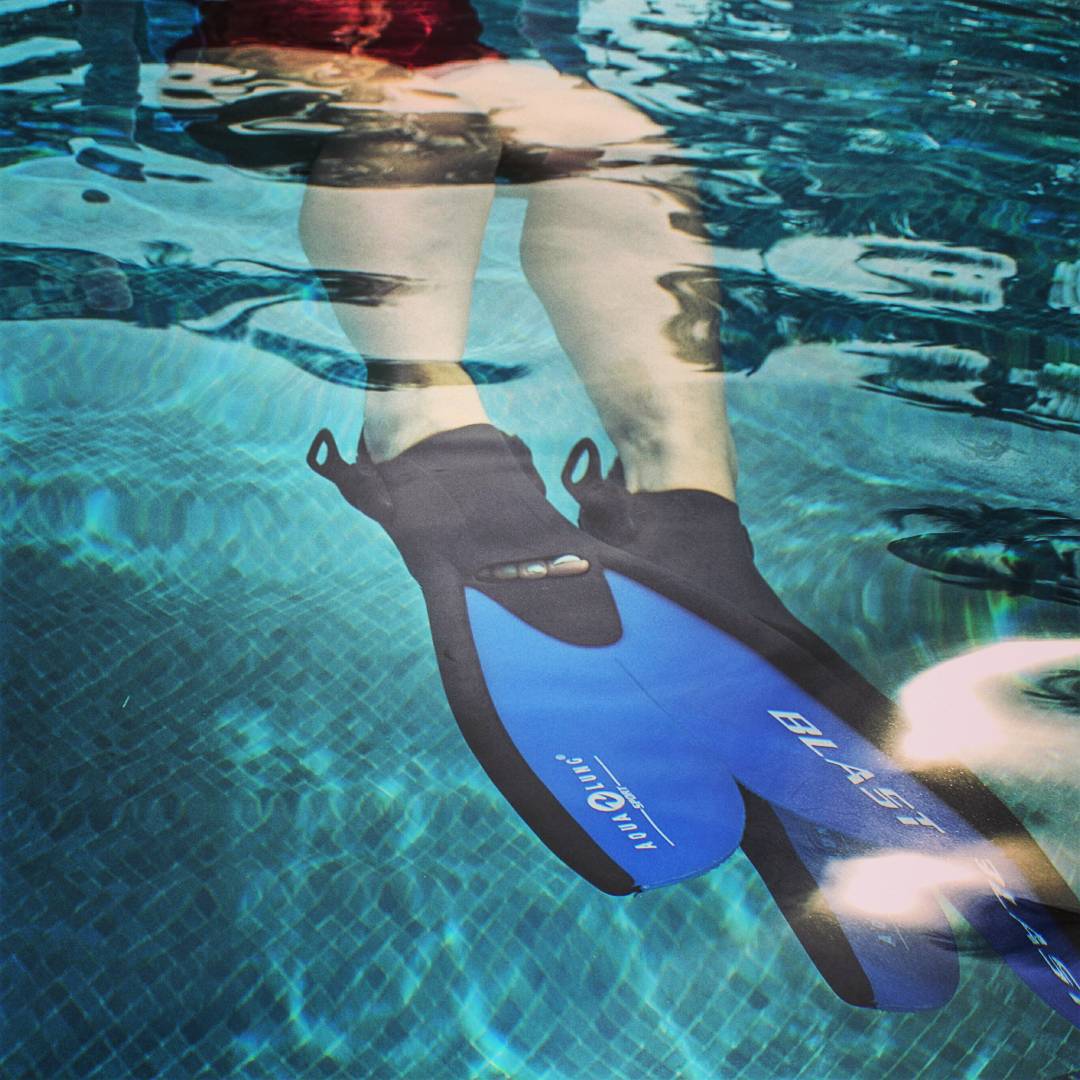 Two legs from the hips down extended under water in a pool with a blue tiled floor. On the person's feet are blue flippers.