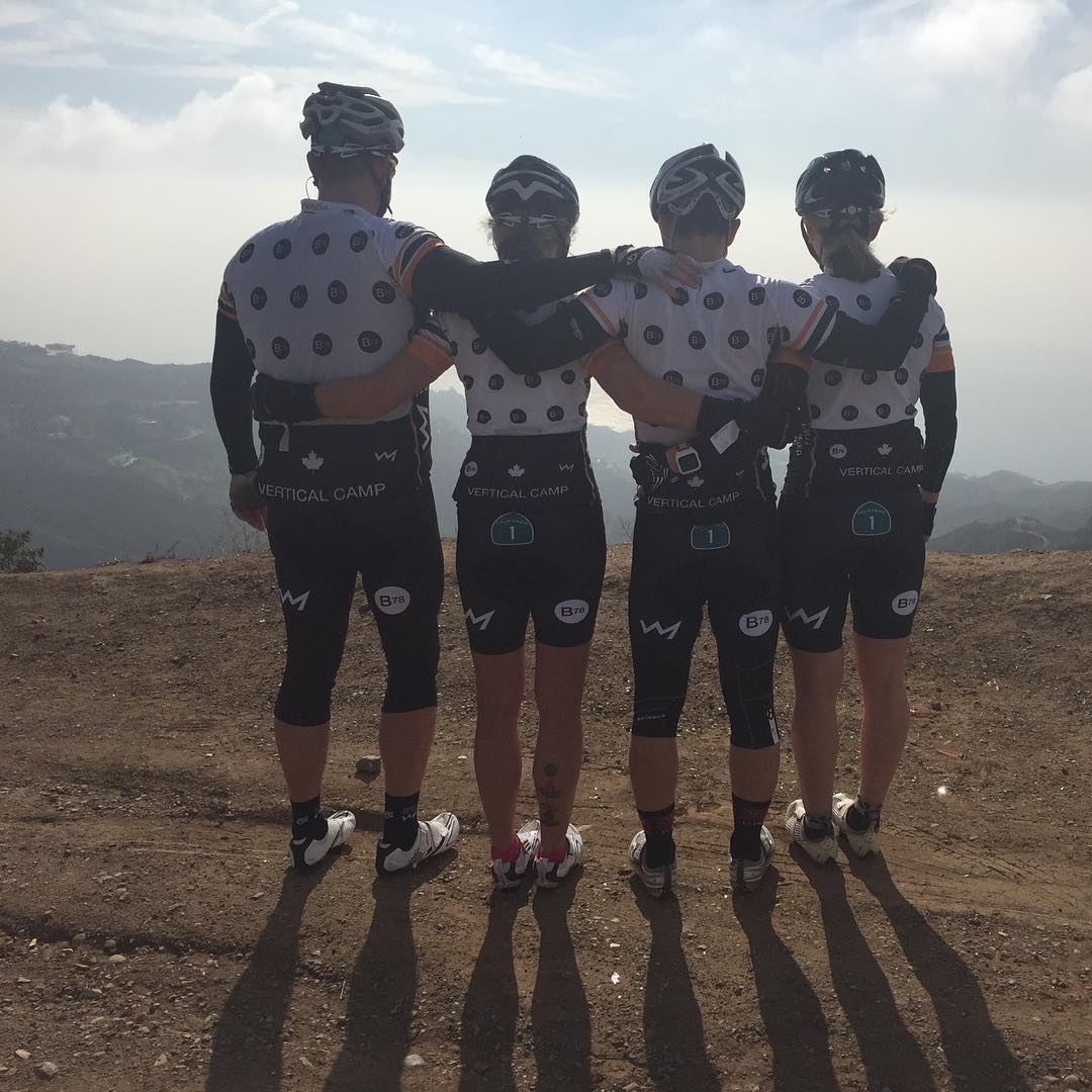 Four people shoulder to shoulder with their arms around each other facing away from the camera to look out over a dry hilly view from a dirt road. The sky is blue with white clouds, and the people are all wearing matching biking outfits with white shirts with black dots and black shorts.