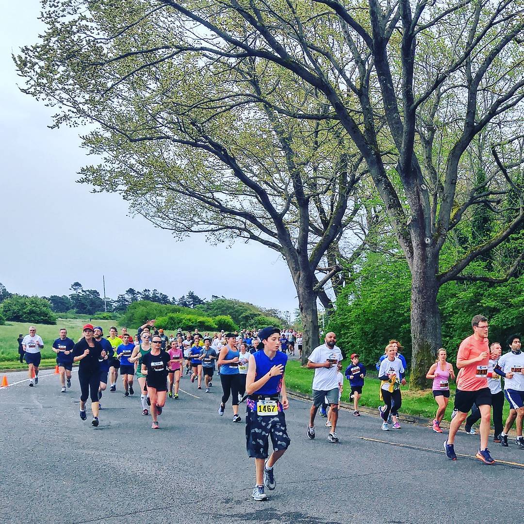 A large group of people running around a bend in the road with race numbers pinned to their stomachs. They are running on the road, and there are green trees and green grass on either side. The sky is a dark, cloudy grey.