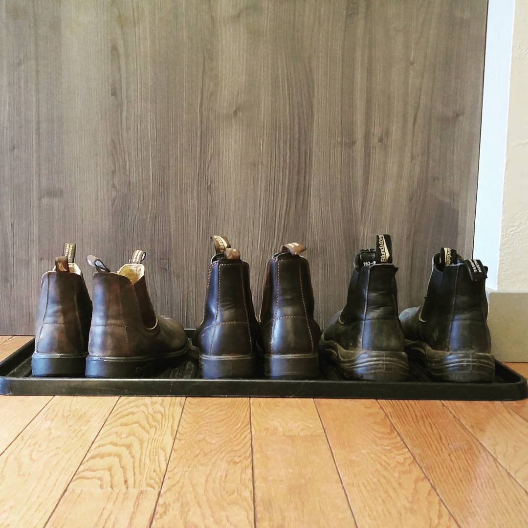 A row of three pairs of brown and black blundstones boots on a black rubber shoe mat on a light hardwood floor. They are in front of a wall that is also wood, though a darker brown.