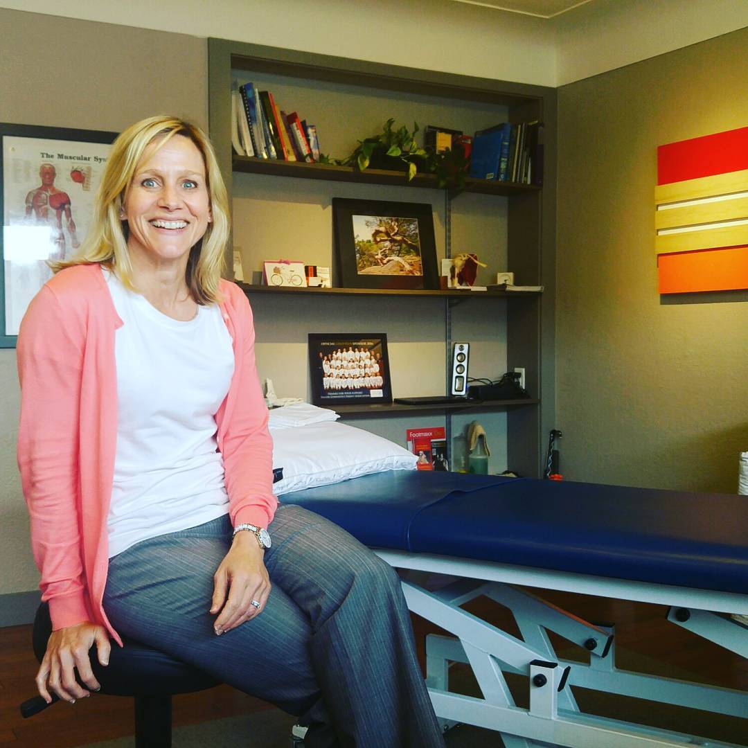 A woman with shoulder length blonde hair, a salmon pink cardigan, blue jeans, and a white tshirt is sitting on a stool in front of a blue treatment bed in an office with brownish green walls. She is smiling widely.