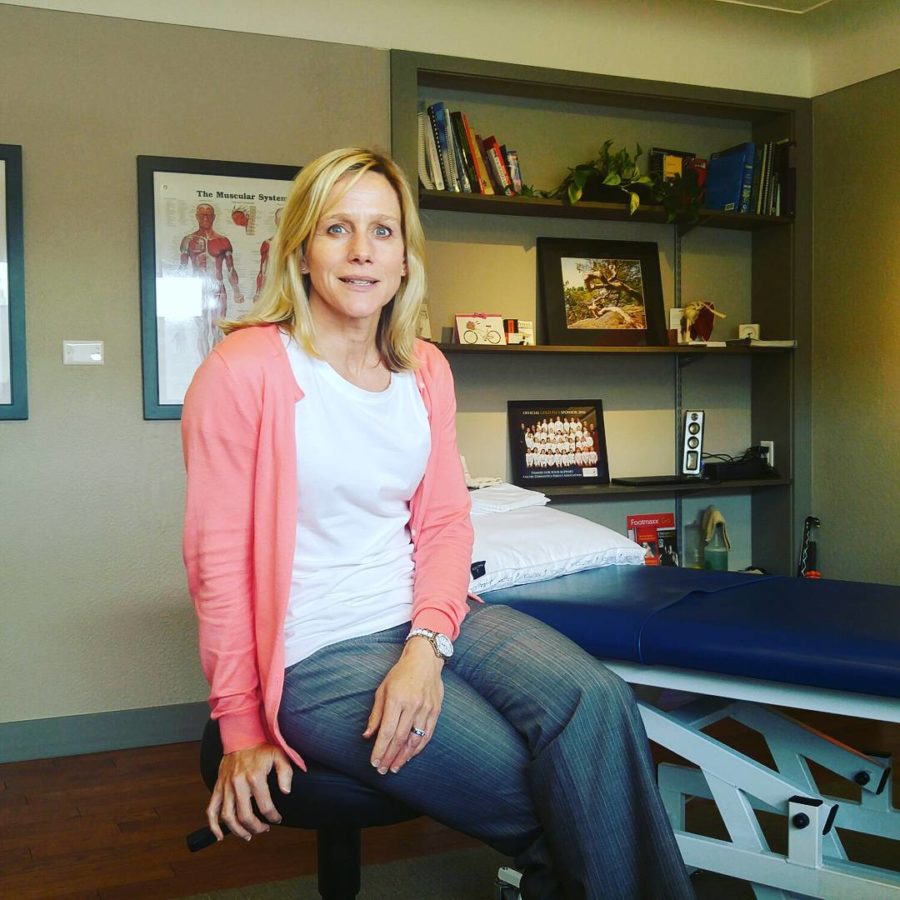 A woman with shoulder length blonde hair, a salmon pink cardigan, blue jeans, and a white tshirt is sitting on a stool in front of a blue treatment bed in an office with brownish green walls. She is smiling only slightly.