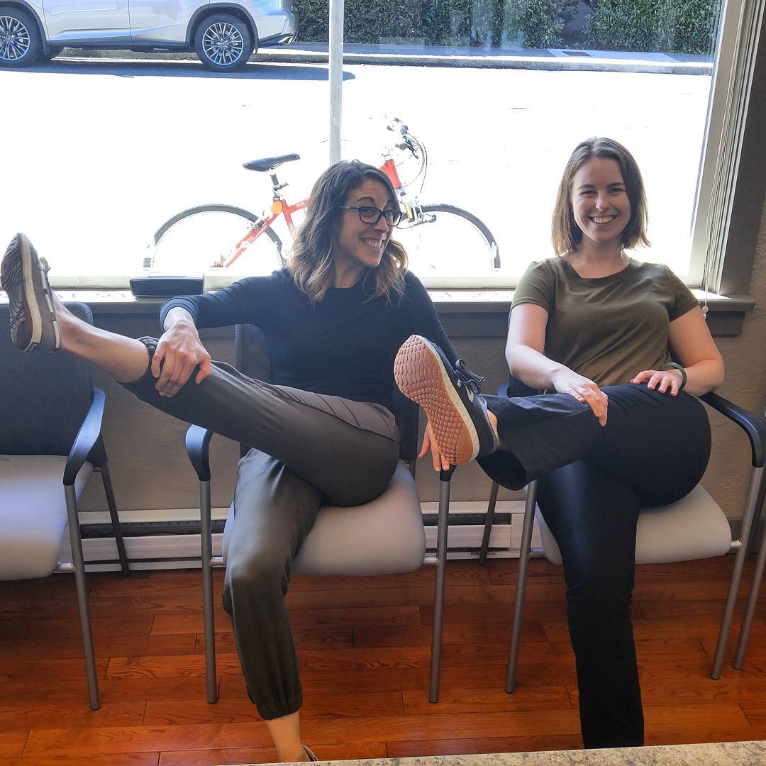 Two women sitting in light brown chairs on reddish hardwood floors. They are both stretching their left legs across their bodies. Both are smiling. It is very bright outside the window.