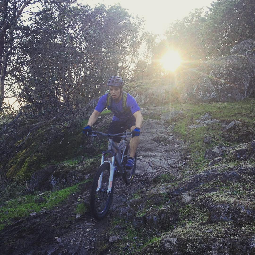 A man in a blue tshirt and a helment biking over damp rocks towards the camera with the sun shining through some thin trees behind him.