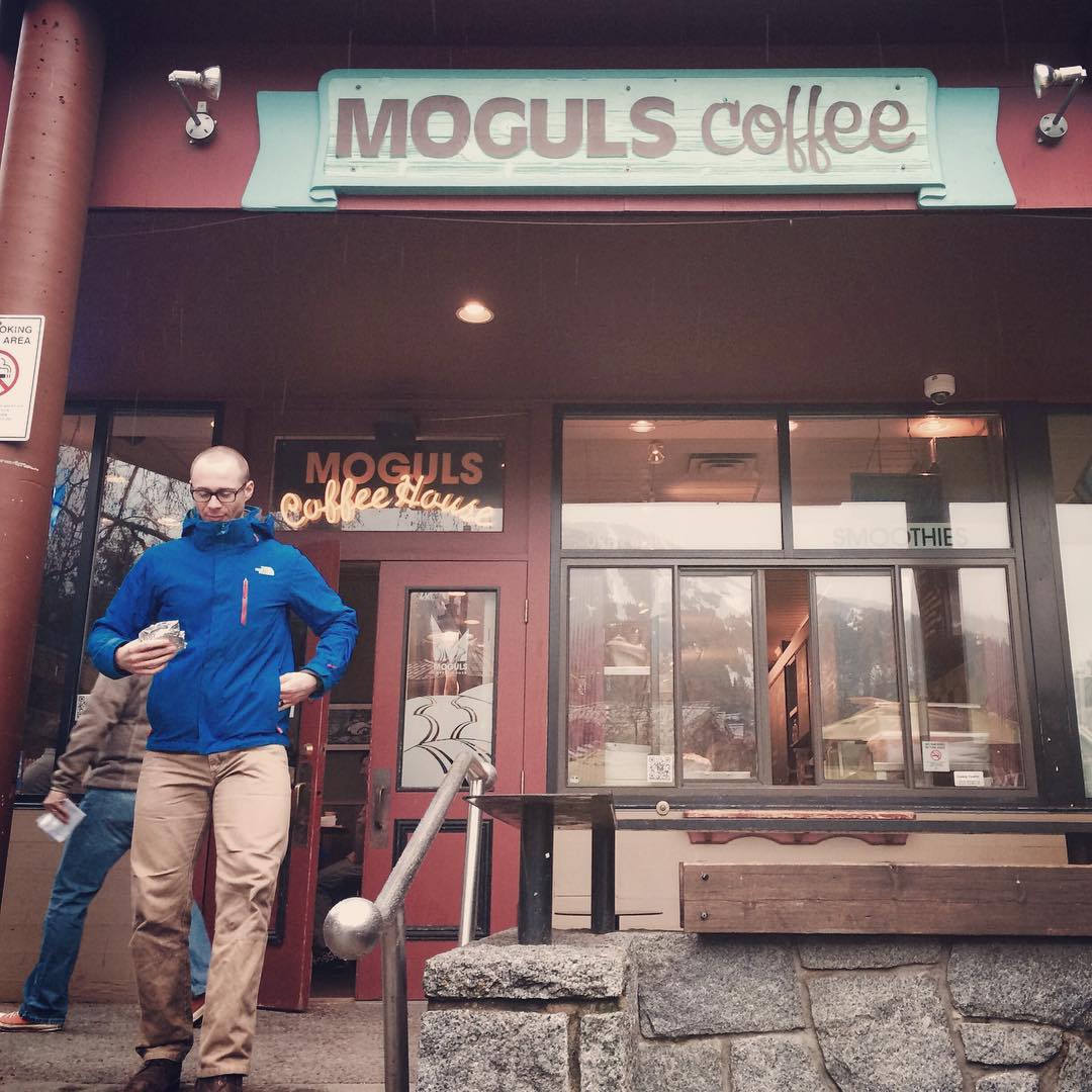 A man in a blue jacket and brown pants carrying a package in his left hand is coming out of a coffee shop with a sign that says "Moguls Coffee."