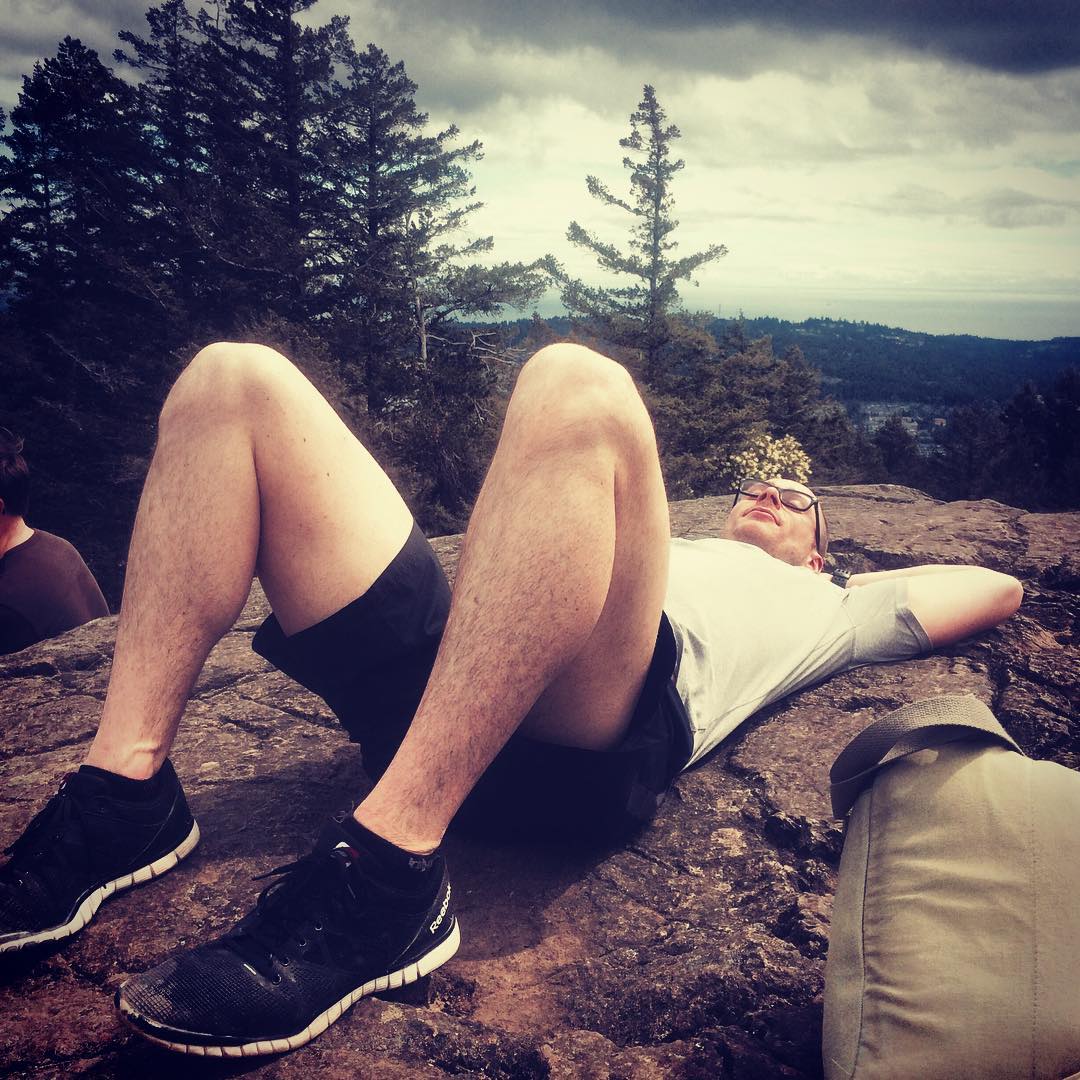 A man lying with his feet towards the camera and arms behind his head is wearing black runners and shorts with a white tshirt. He is lying on rocky ground with evergreens in the distance. The sky is grey and cloudy.