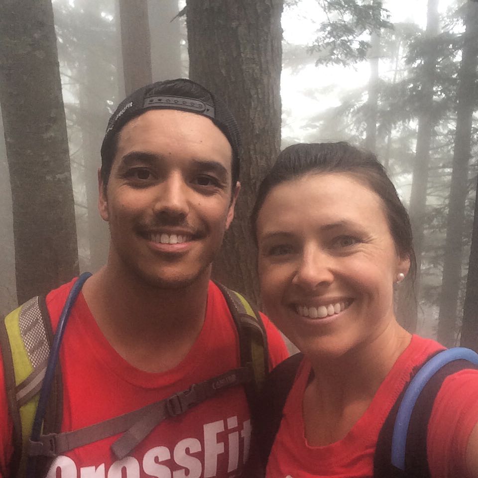 A man on the left and a woman on the right both wearing backpacks and red tshirts. They are shoulder to shoulder and smiling widely and the camera. Tall evergreen trees and fog are behind them.