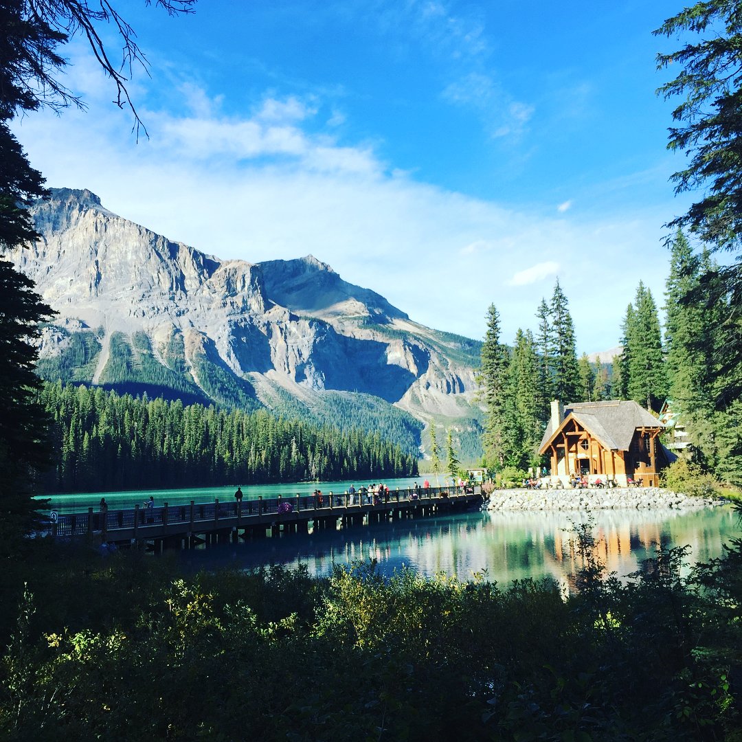 A view of an emerald coloured lake with a wooden cabin in the distance right, and mountains rising to the left. It is framed by evergreen trees and a bright blue sky.
