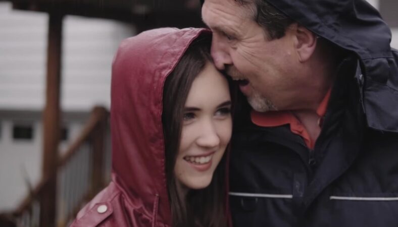 A father (right) and daughter (left) with their heads together smiling widely in the rain.