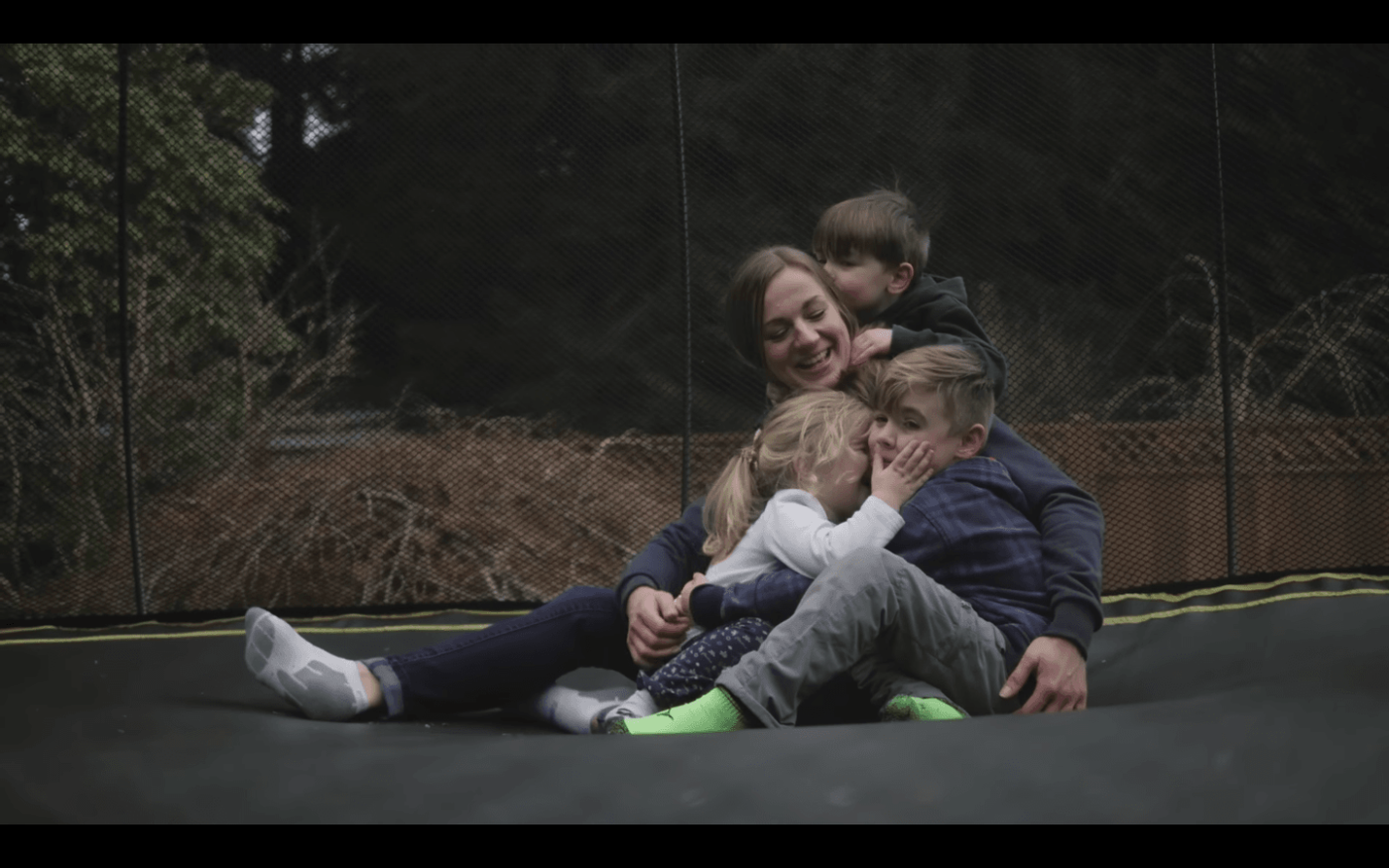 A woman sitting on a trampoline smiling widely with three toddlers climbing all over her and kissing her.