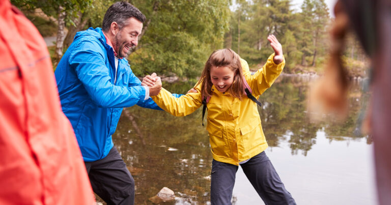 A father reaches out to help his daughter balance on a hike by water.