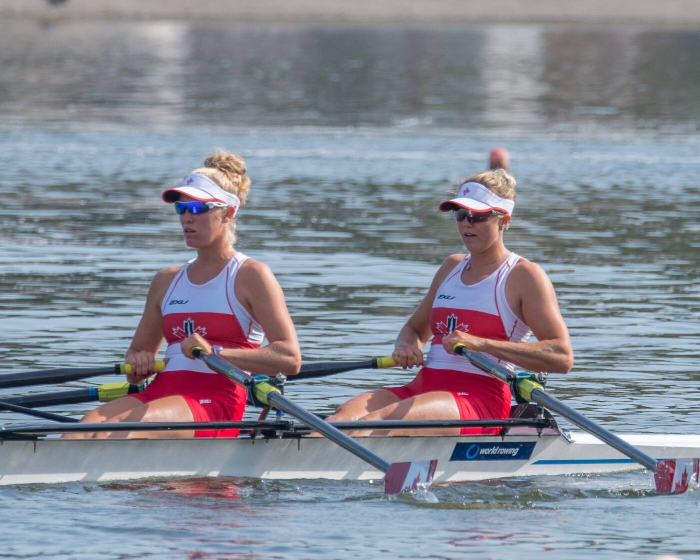 Two women wearing red and white uniforms and visors rigorously rowing on calm water.