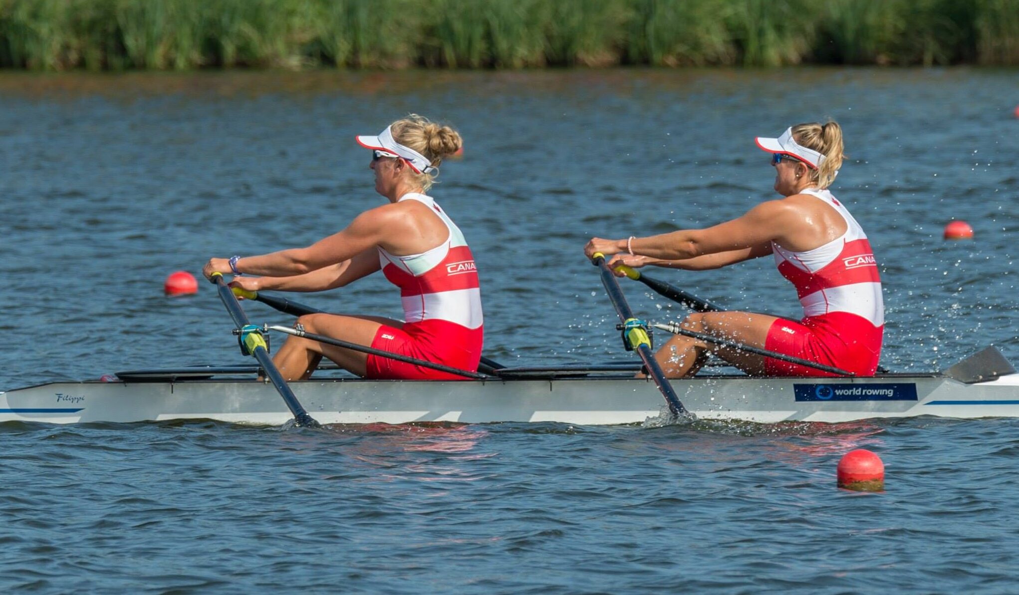 Two women wearing red and white uniforms rigorously rowing on calm water with tall grass in the background.