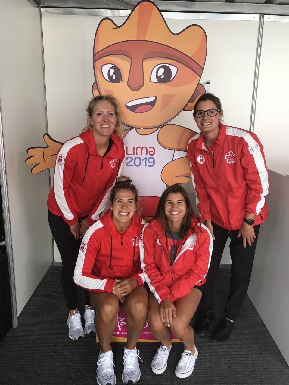 Four women in matching red and white team jackets pose in front of a cut-out of the Lima Pan Am Games 2019 mascot smiling.