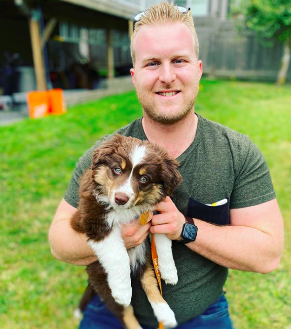 Connor kneels in the grass and smiles at the camera with a puppy in his arms. The puppy looks like a brown and white aussie shepherd.