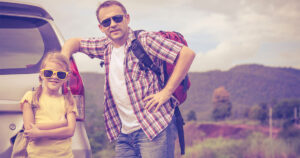 A father and daughter stand posing against a van with a dry landscape behind them wearing sunglasses and backpacks.