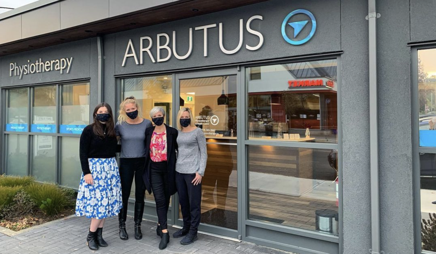 Four masked women pose in front of the main doors to the new Arbutus Clinic to celebrate one year on Oak Bay Avenue