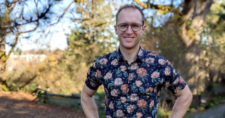 Maciek stands with his arms on his hips in a park, wearing a dark blue shirt that has roses on it.