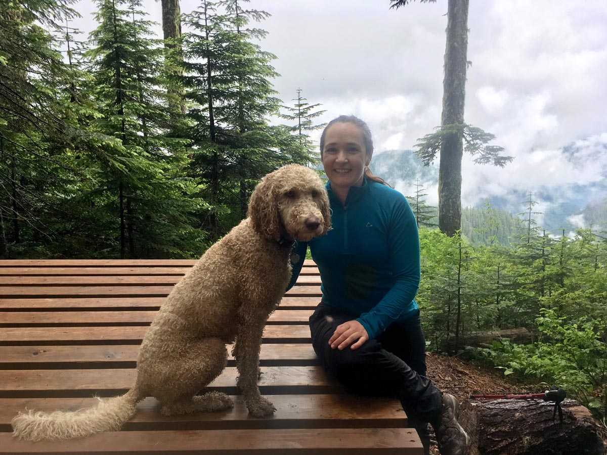 Jacqueline McAllister, physiotherapist, sits on a bench with Sadie her dog in the forests of Vancouver Island on a cloudy day.