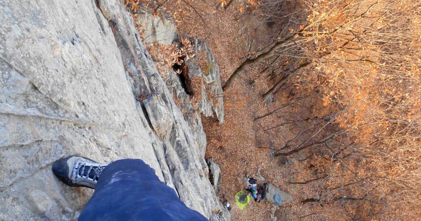 The climbers takes a photo looking down the cliff face with the people seen down below and the climber's foot in the picture.