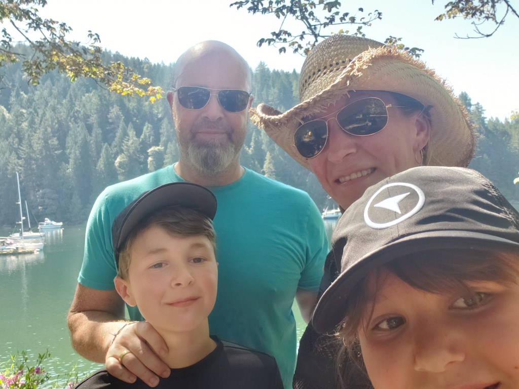 Natasha Watson wears sunglasses and a straw hat, smiling alongside her husband who is also wearing sunglasses, and her two children as they take a selfie in front of a lake with trees in the background.