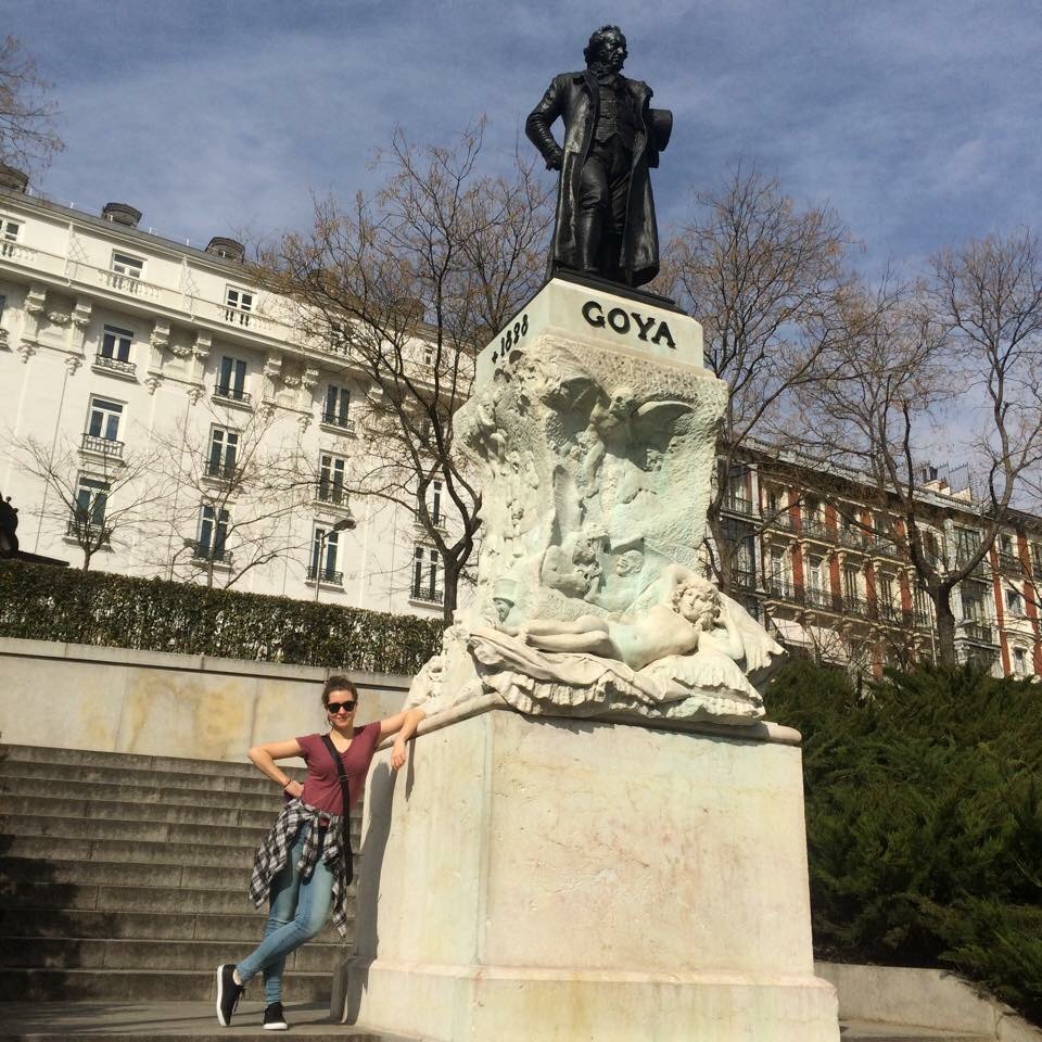 Zoë Stocker stands wearing sunglasses and a mauve t-shirt, with her arm propped on a statue of Goya.