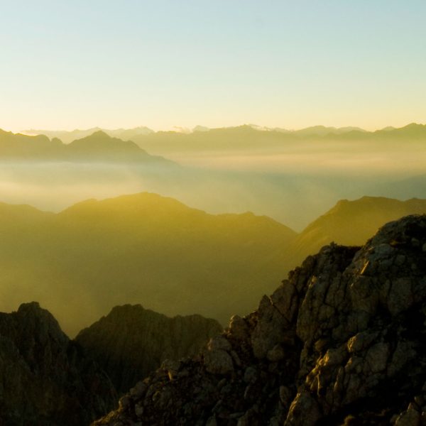 A person stands atop a mountain range looking out over the clouds and sunset light.
