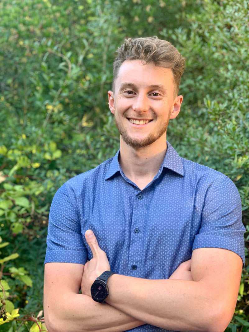 Joseph Rector, MScPT, CSEP-CEP, physiotherapist, smiles and crosses his arm, wearing a blue collared shirt and standing in front a tall green bushy backdrop.