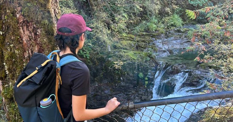 Hatty peers over a fence at the waterfall below, while wearing a maroon baseball cap and black backpack.