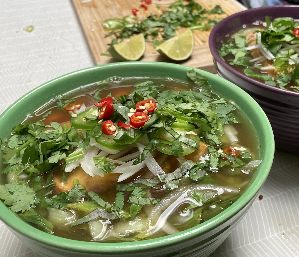 A green bowl full of southeast Asian-style noodle soup full of red chilies, cilantro, and limes.