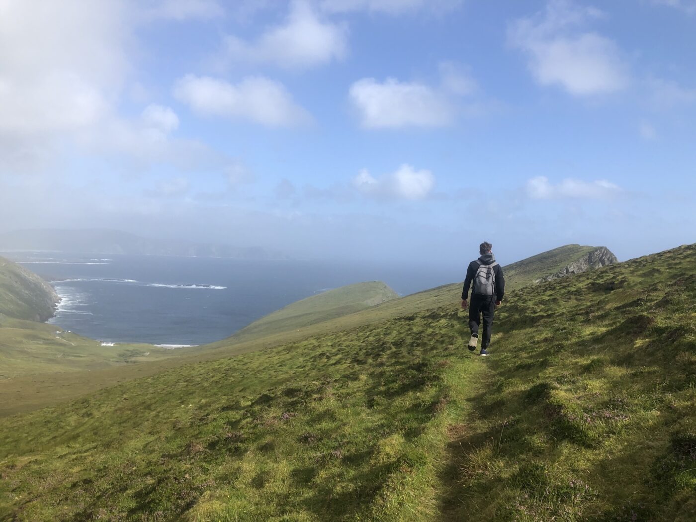 Joseph Rector hikes a grassy hillside along the shore of Ireland's coastline with the ocean in the distance.