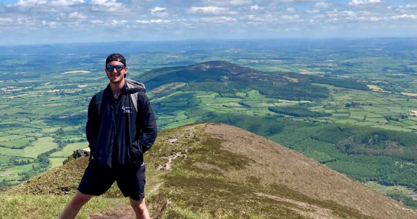Joseph Rector stands on top of a mountain in Ireland, wearing sunglasses and a backward baseball cap, with rolling green hills in the background.