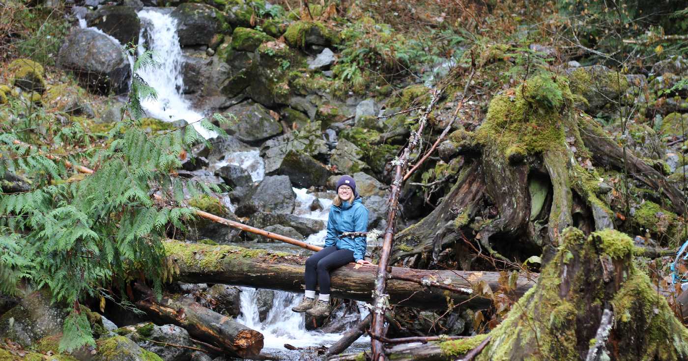 Madi Campbell sits on a fallen tree over a waterfall and rushing river in a cedar forest.