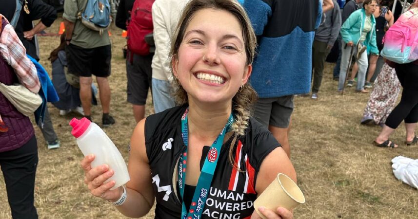 Kezia Dawes smiles at the camera after a race, wearing a medal while holding a plastic water bottle and an empty coffee cup.