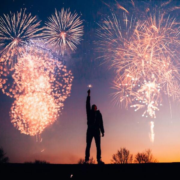 A person holds up a lit sparkler while fireworks burst behind them at sunset.