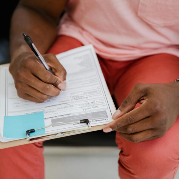 A person of colour wearing a pink shirt and salmon coloured pants writes on a clipboard.