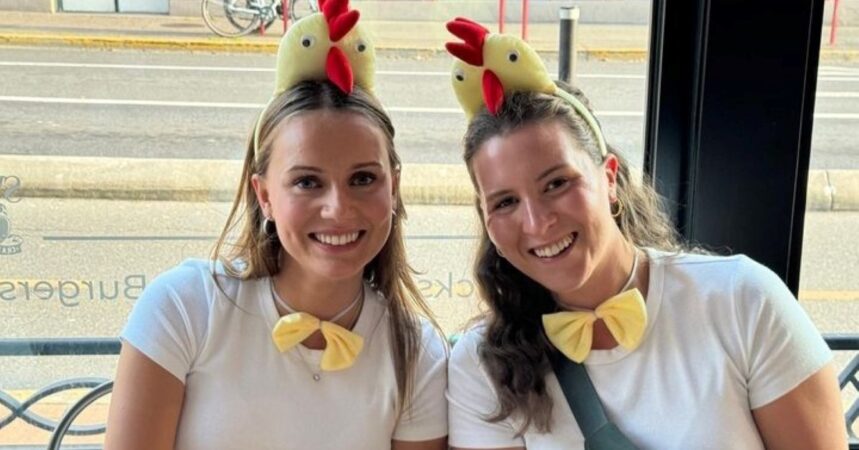 Two people sit together smiling at the camera and wearing chicken costumes with chicken headbands, yellow bow ties, and yellow tutus.