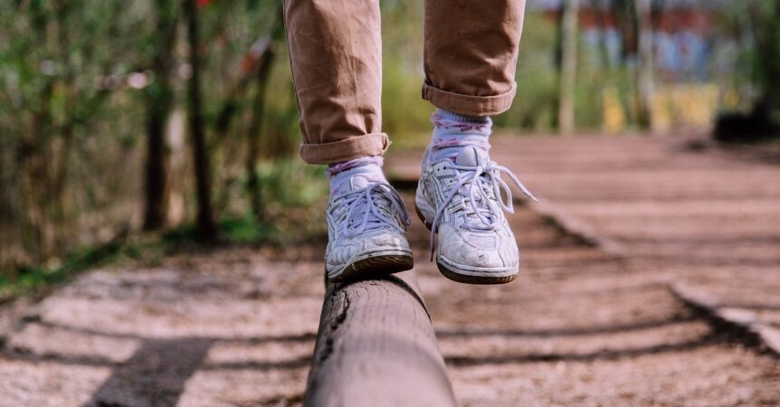 A close-up of someone's feet balancing on a fallen log. They are wearing white sneakers and brown trousers.