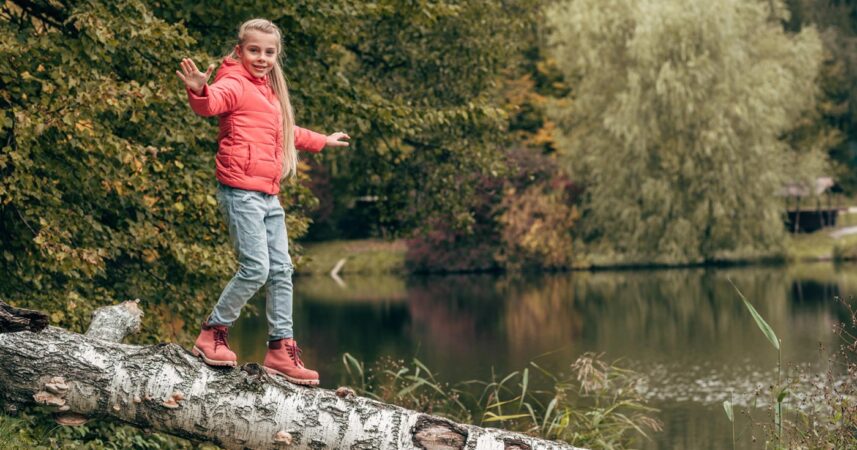 A girl wearing a peach coat and boots walks along a log over a lake.